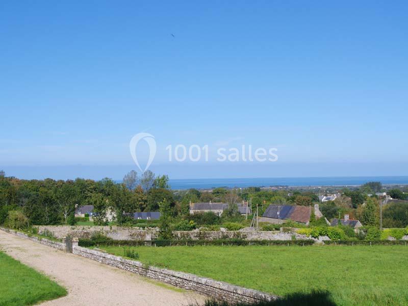 Vue d'un village avec des maisons entourées de verdure, un chemin de terre et la mer visible à l'horizon sous un ciel bleu.