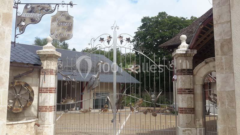 Portail en fer forgé décoré de motifs d'oiseaux, entouré de piliers en pierre, devant un bâtiment à toit en ardoise.