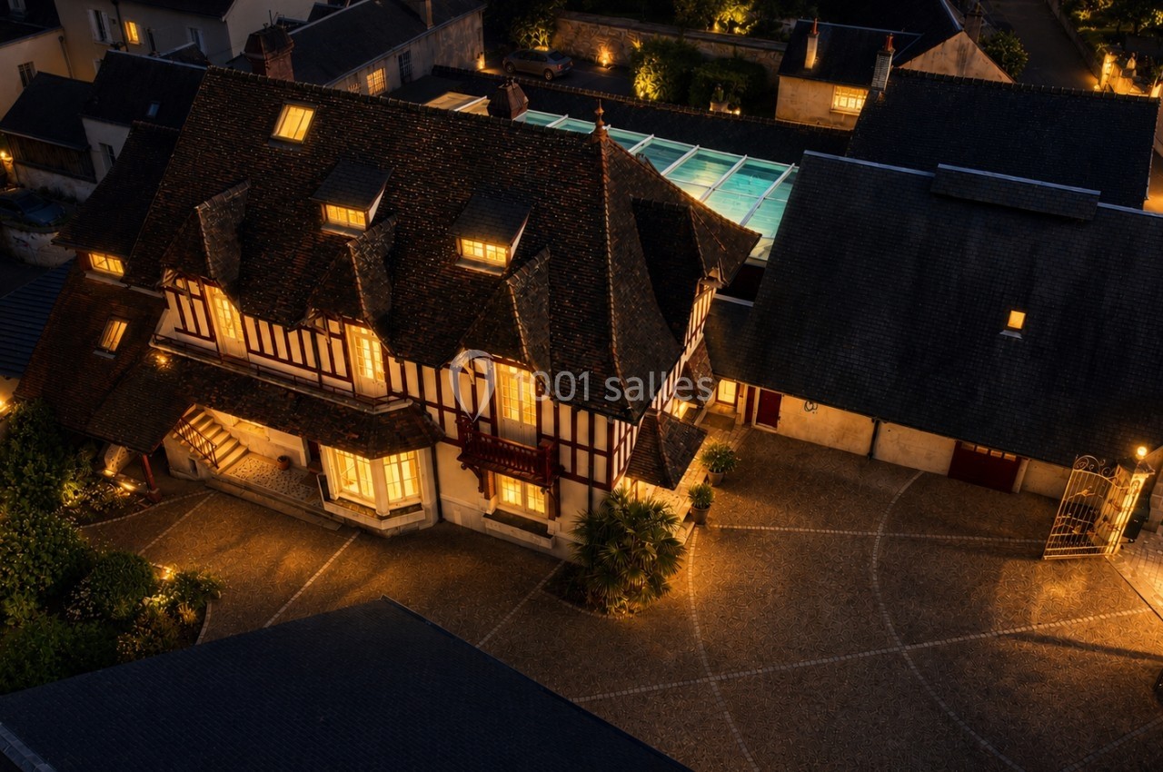 Vue aérienne nocturne d'une maison à colombages éclairée, entourée de bâtiments et d'une cour pavée.