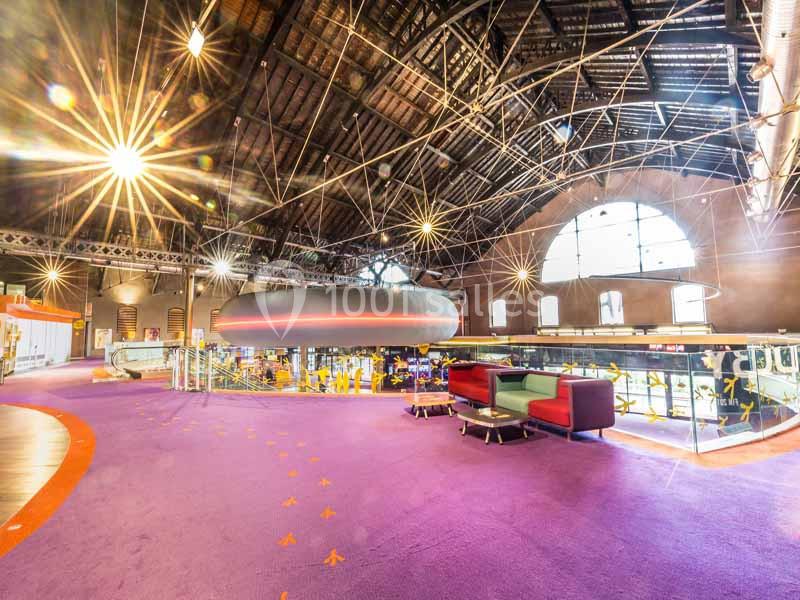 Intérieur d'un bâtiment avec un plafond voûté, un sol violet, des canapés colorés et une structure suspendue moderne.