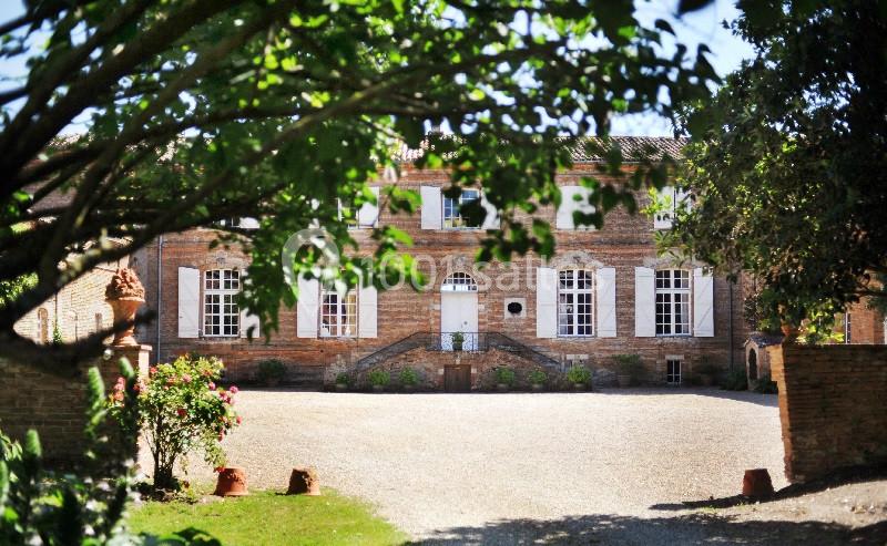 Façade d'une maison en briques rouges avec volets blancs, entourée d'arbres et donnant sur une cour gravillonnée.