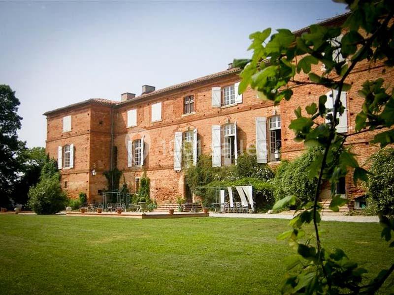 Façade d'un grand bâtiment en briques rouges avec volets blancs, entouré de verdure et d'une pelouse bien entretenue.
