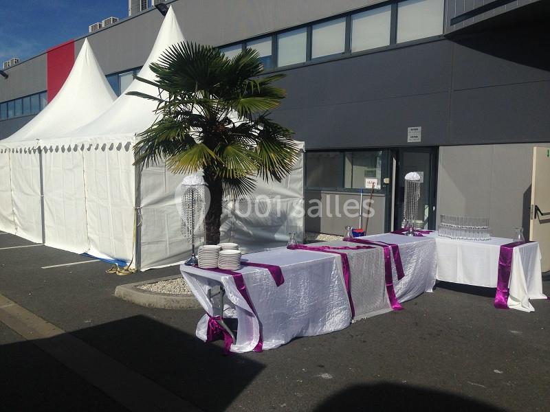 Tentes blanches et tables décorées de nappes blanches et rubans violets, installées en extérieur près d'un bâtiment.