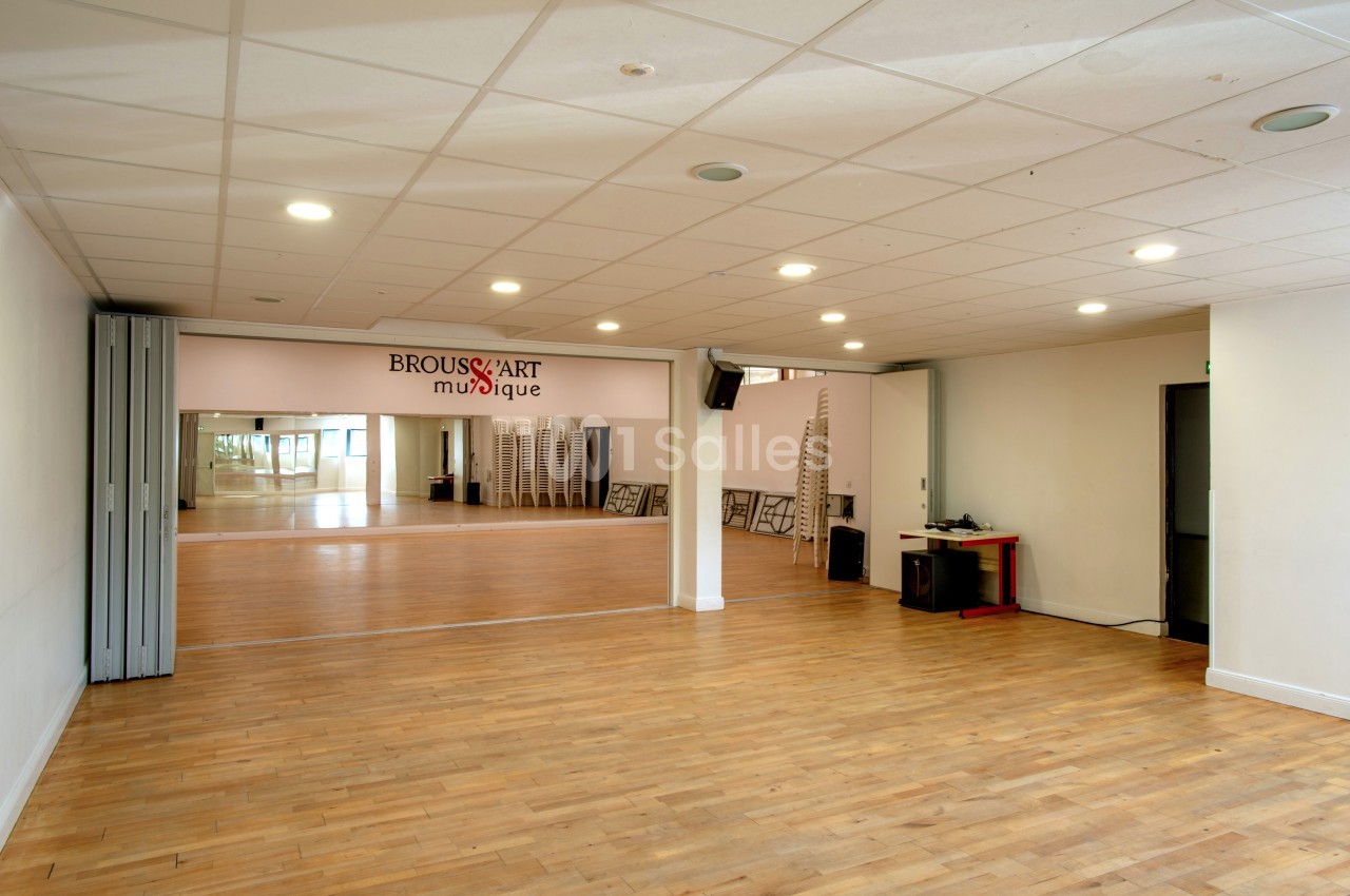 Salle lumineuse avec parquet en bois, miroirs sur un mur, chaises empilées et équipement audio dans un coin.