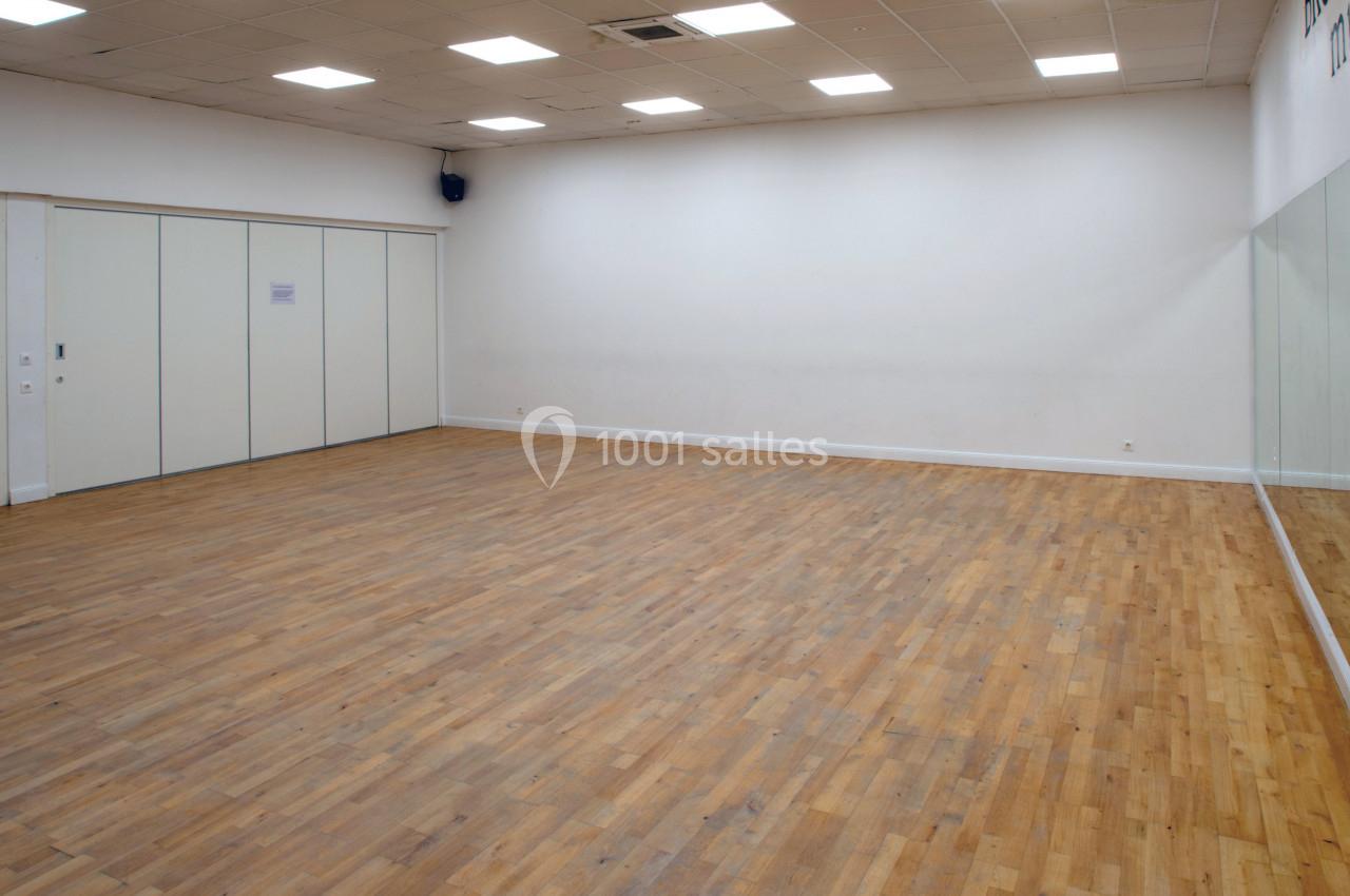 Salle vide avec parquet en bois, murs blancs, plafond à dalles lumineuses et grand miroir sur un côté.