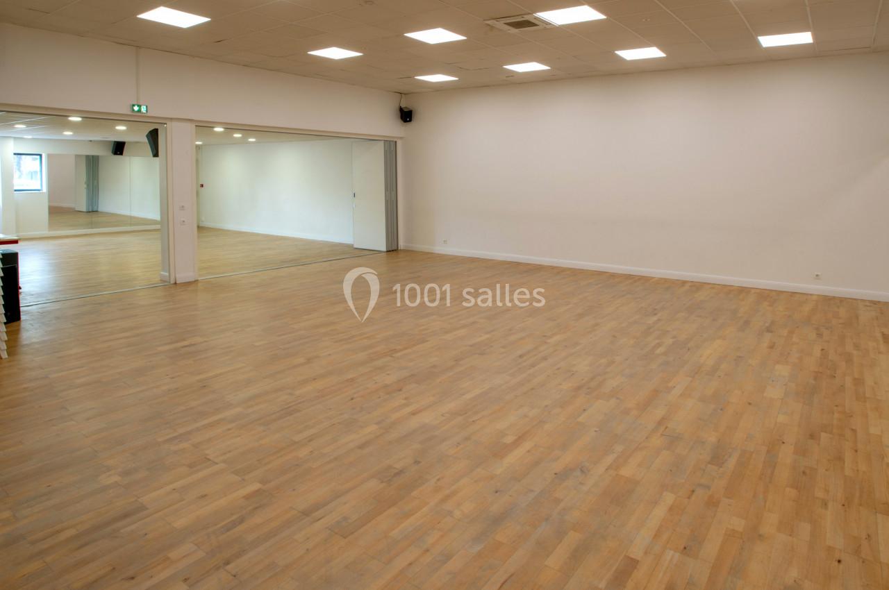 Salle vide avec parquet en bois, murs blancs, miroirs sur un côté et chaises empilées dans un coin.