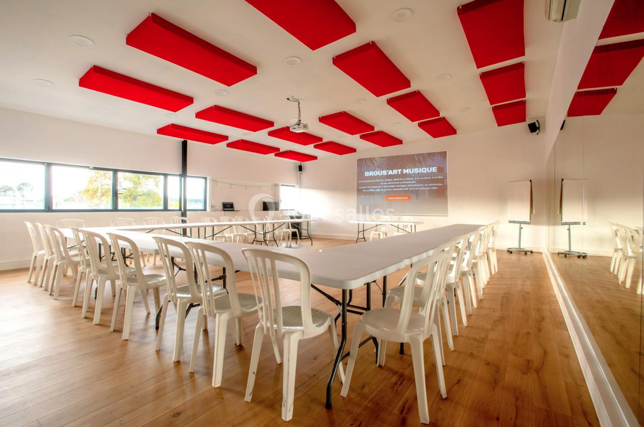 Salle lumineuse avec tables blanches, chaises en plastique, panneaux acoustiques rouges au plafond et écran de projection.