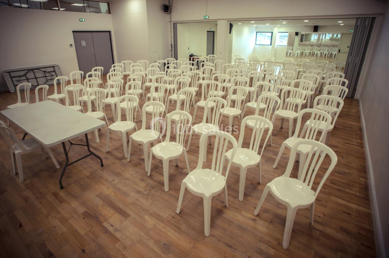 Salle avec parquet clair, rangées de chaises blanches en plastique et une table rectangulaire à l'avant.