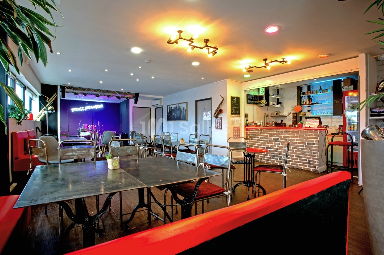 Salle de restaurant avec tables en métal, bar en briques, éclairage chaleureux et scène équipée en arrière-plan.