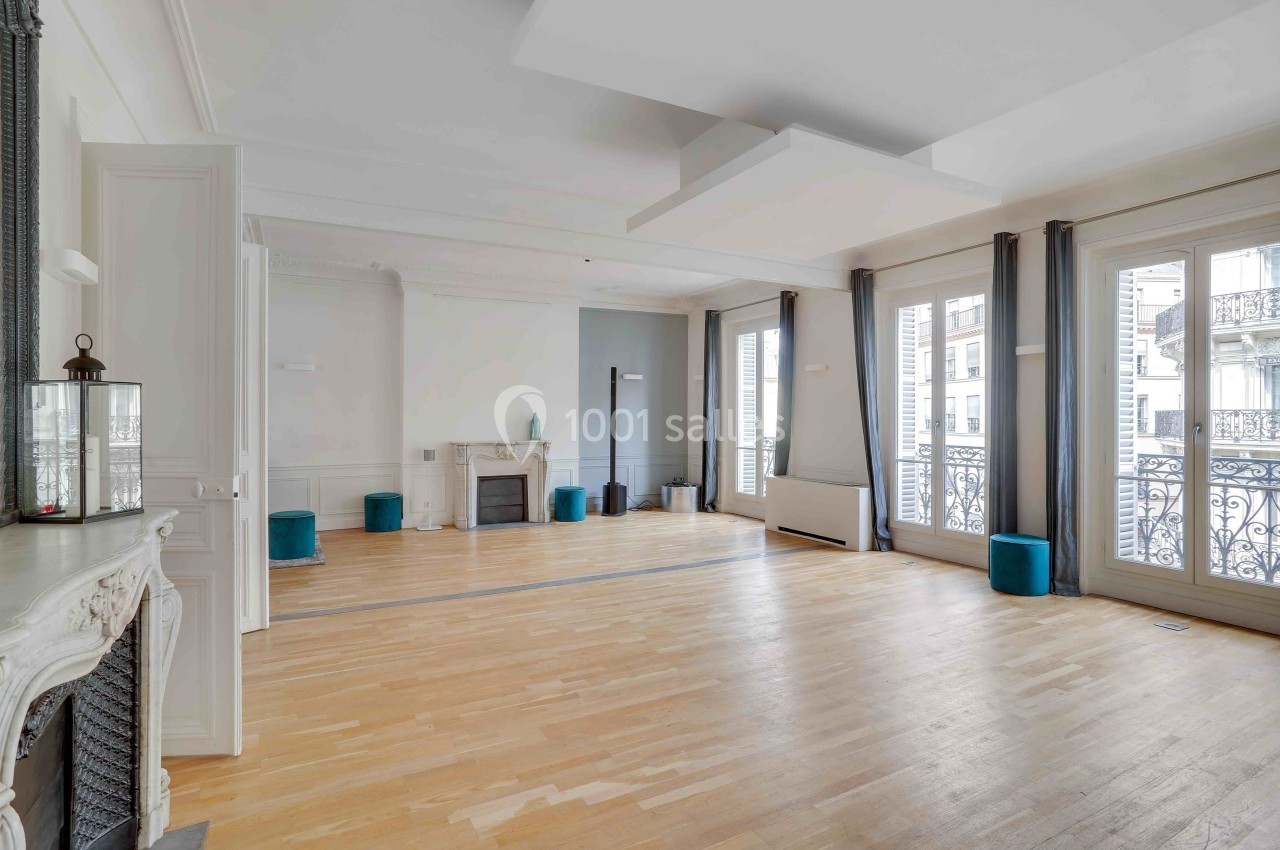 Salle lumineuse avec parquet clair, cheminée décorative, grandes fenêtres et rideaux gris donnant sur un balcon en fer forgé.