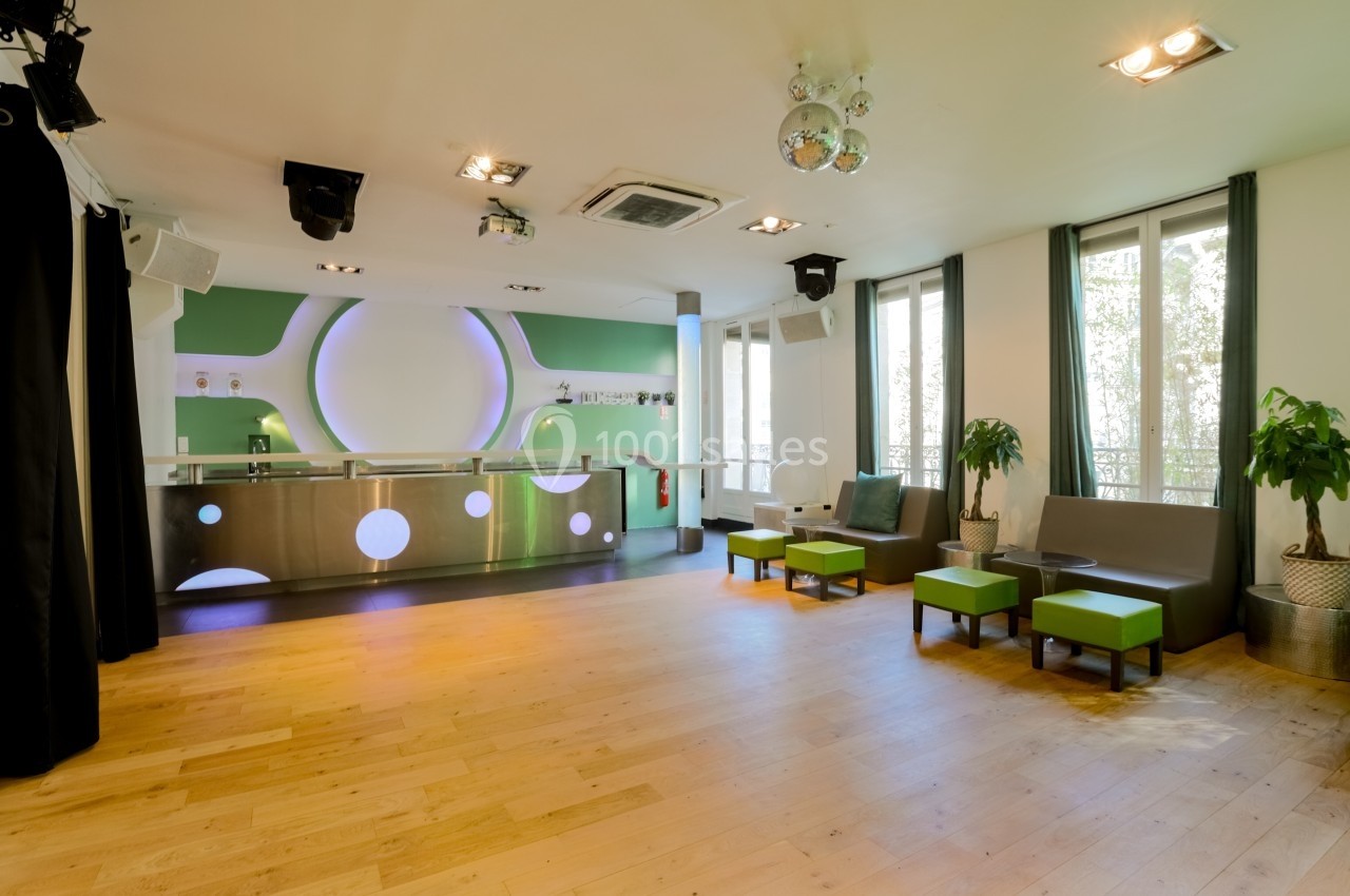 Salle lumineuse avec parquet, comptoir moderne vert et blanc, sièges verts, grandes fenêtres et plantes décoratives.