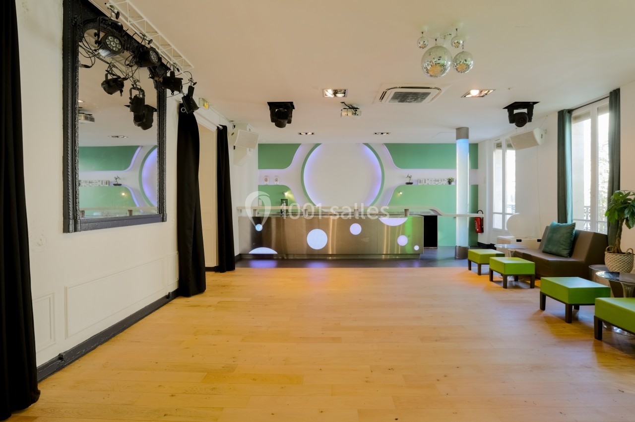 Salle lumineuse avec parquet clair, comptoir moderne en métal, murs verts et sièges verts près des fenêtres.