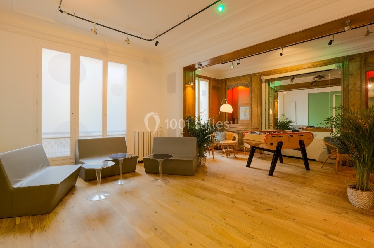 Salle lumineuse avec parquet, canapés gris, baby-foot, plantes et grandes fenêtres laissant entrer la lumière naturelle.