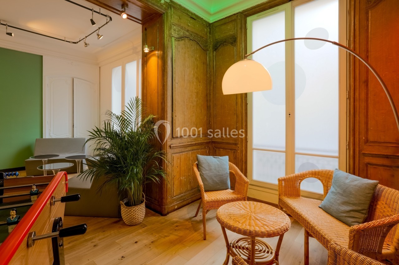 Espace intérieur lumineux avec fauteuils en rotin, table basse, plante verte et baby-foot, encadré de boiseries.