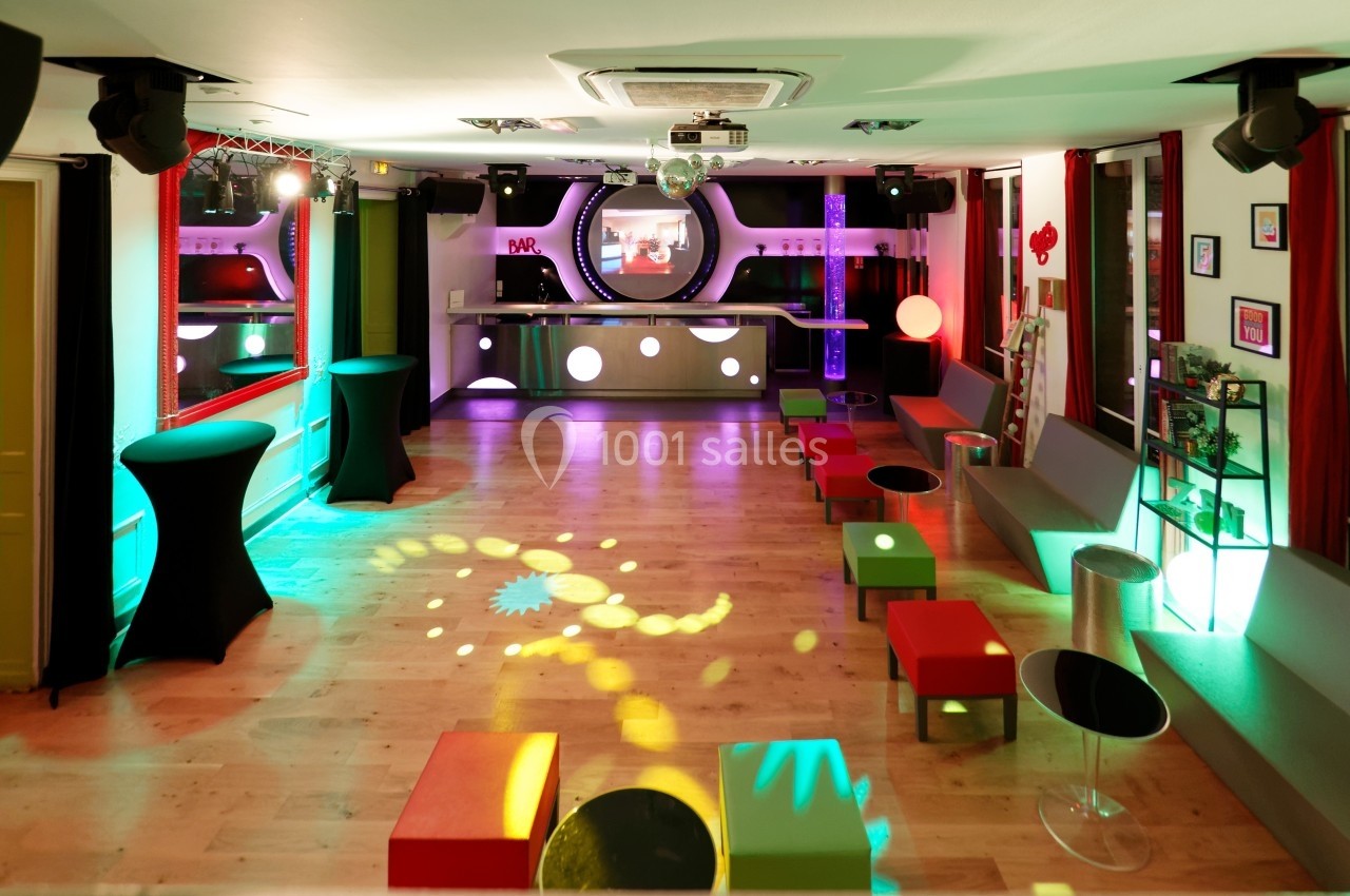 Salle colorée avec éclairages festifs, bar en arrière-plan, sièges modernes et piste de danse en bois clair.