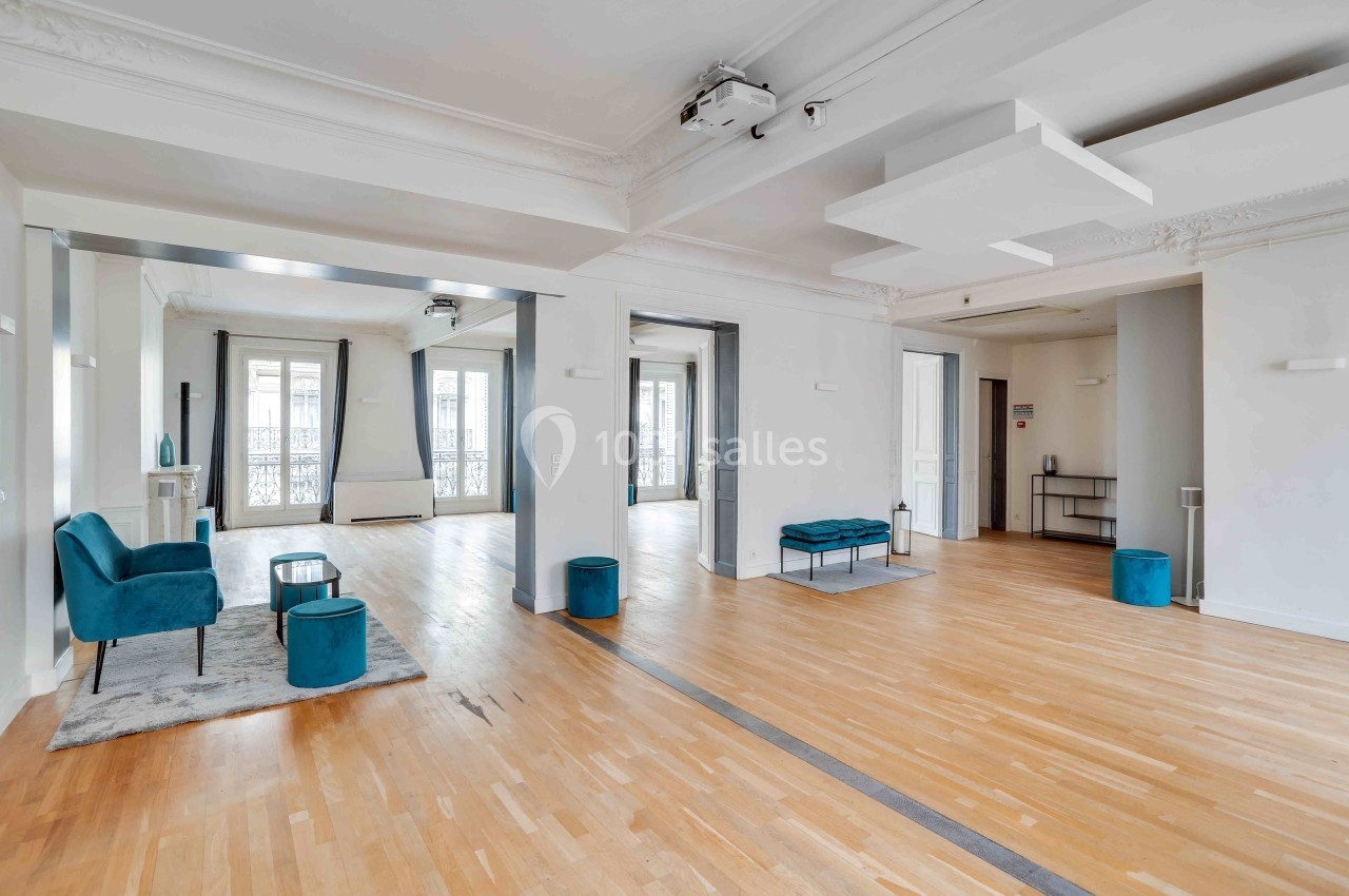 Salle lumineuse avec parquet clair, fauteuils bleus, grandes fenêtres et éléments de décoration modernes.