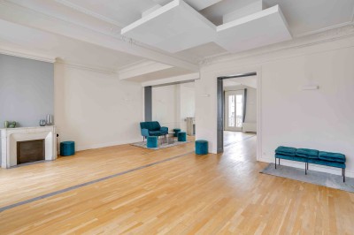 Salon lumineux avec parquet en bois clair, cheminée, fauteuils bleus et moulures au plafond.