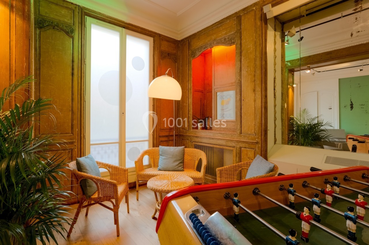 Salle conviviale avec baby-foot, fauteuils en rotin, plantes et éclairage chaleureux près d'une fenêtre.