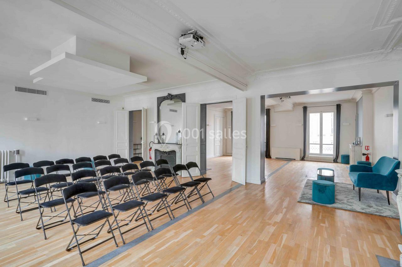 Salle lumineuse avec parquet, rangées de chaises noires, cheminée décorative et espace détente avec fauteuils.