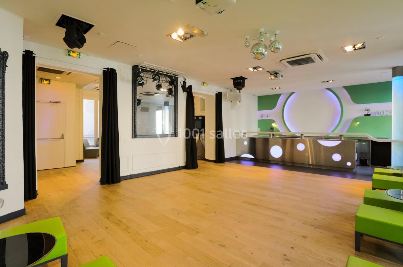 Salle lumineuse avec parquet en bois clair, bar moderne au fond et sièges verts le long des murs.