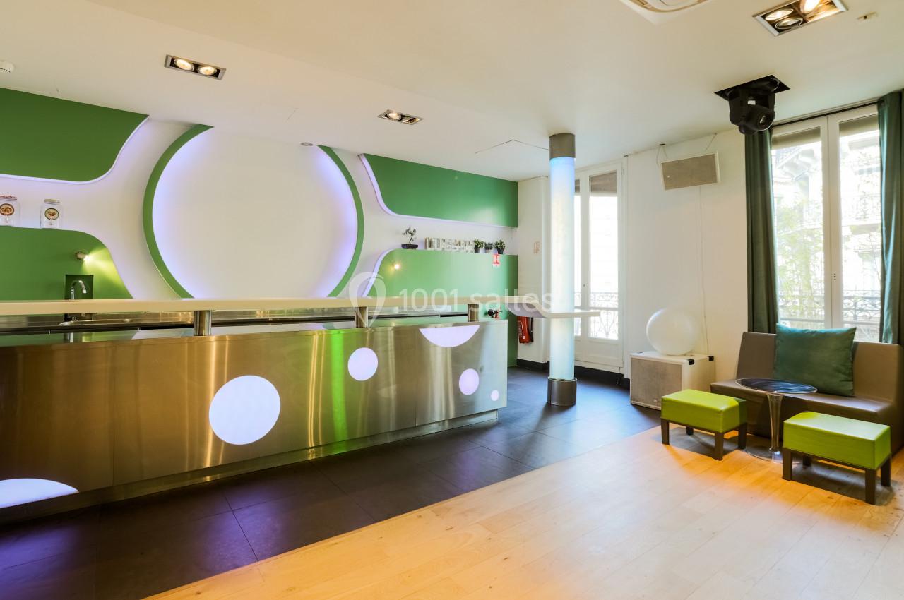 Intérieur lumineux d'un espace moderne avec comptoir métallique, murs verts et blancs, et coin salon avec fauteuils.