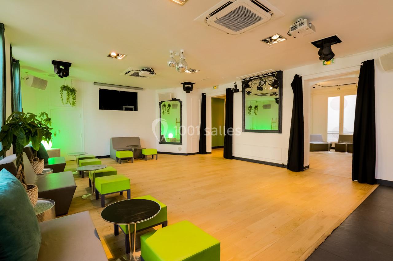 Salle lumineuse avec parquet clair, fauteuils verts et gris, plantes décoratives et éclairage d'ambiance vert.