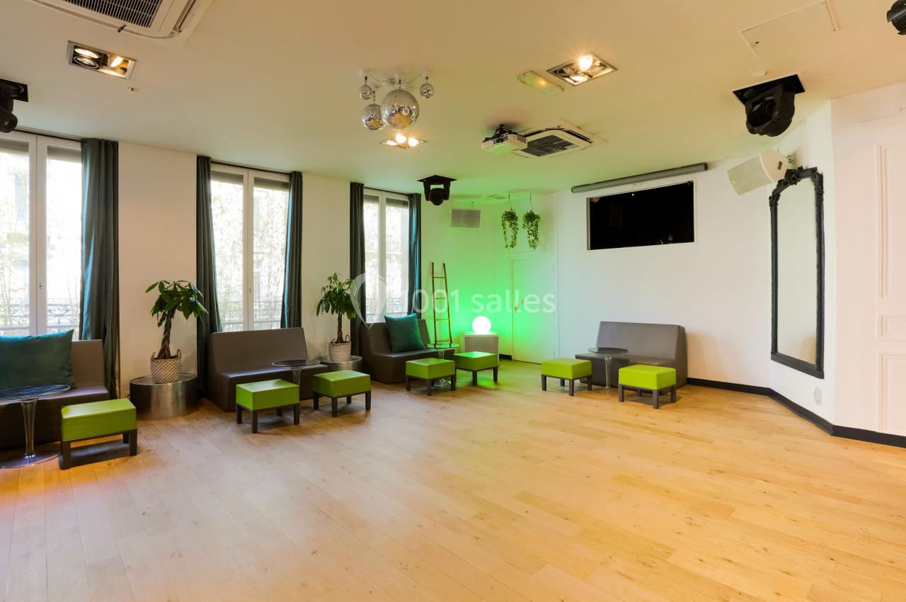 Salle lumineuse avec parquet clair, sièges gris et verts, grandes fenêtres, plantes et éclairage décoratif.