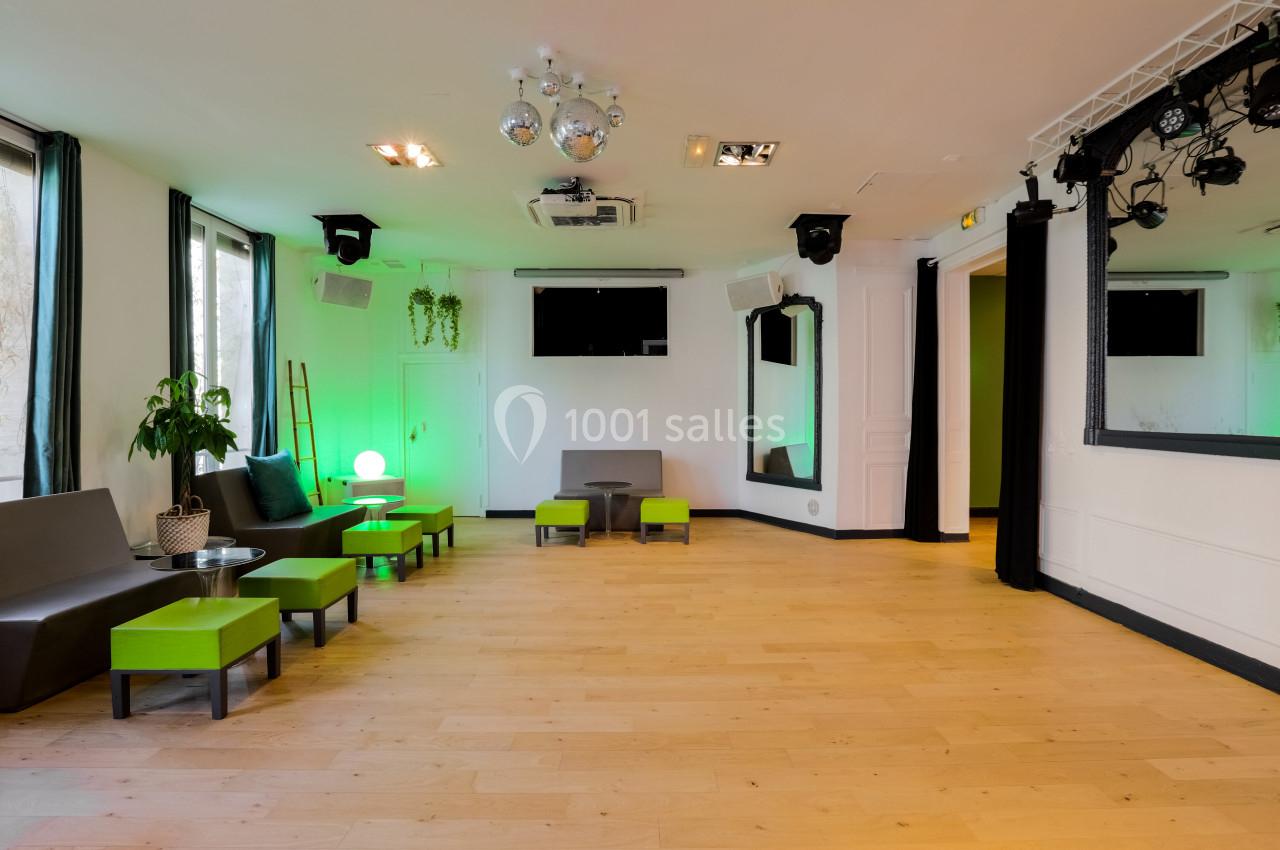 Salle lumineuse avec parquet clair, mobilier moderne vert et gris, éclairage décoratif et scène équipée de projecteurs.