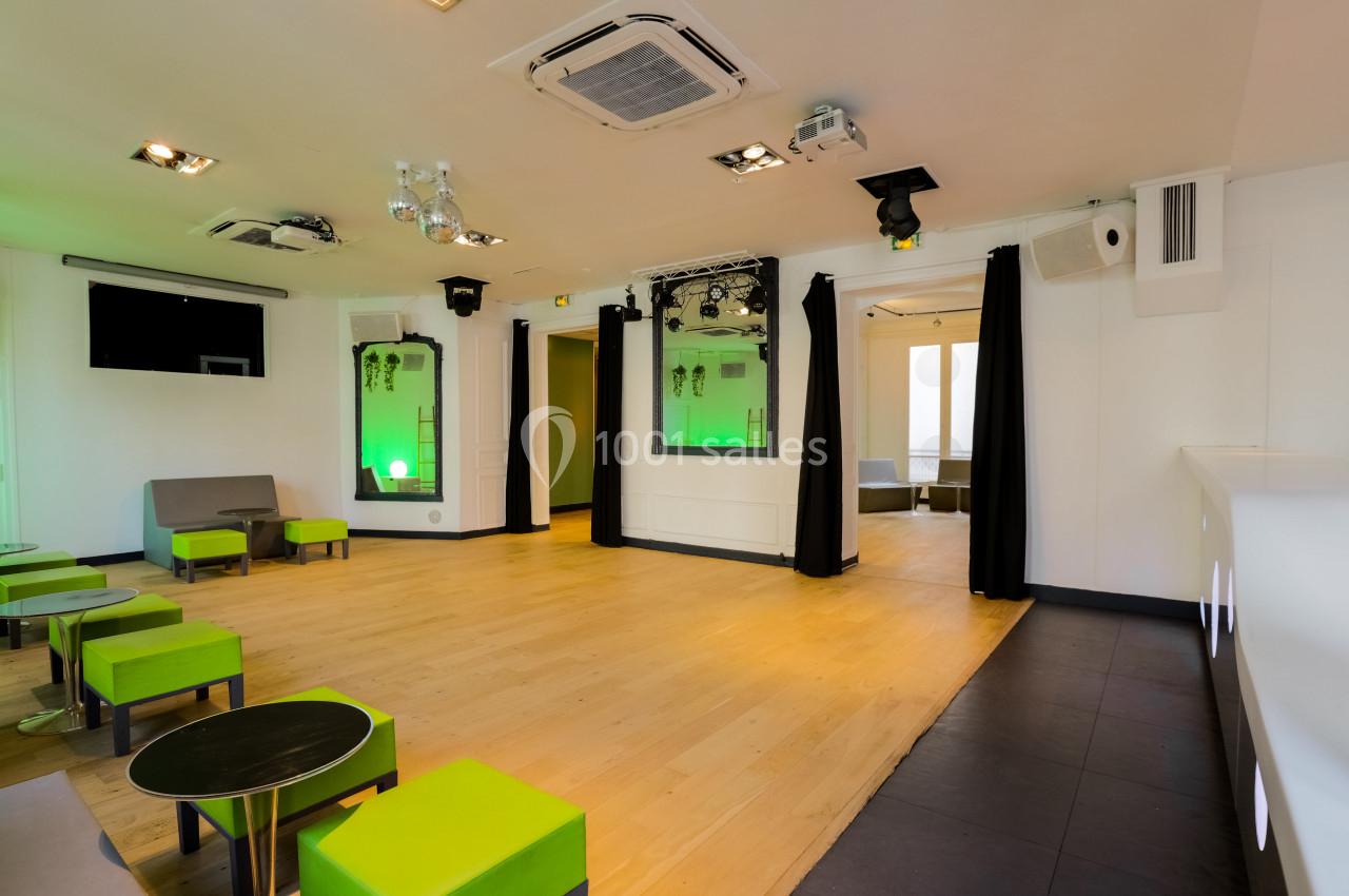 Salle lumineuse avec parquet, sièges verts, tables basses, rideaux noirs et éclairage moderne.