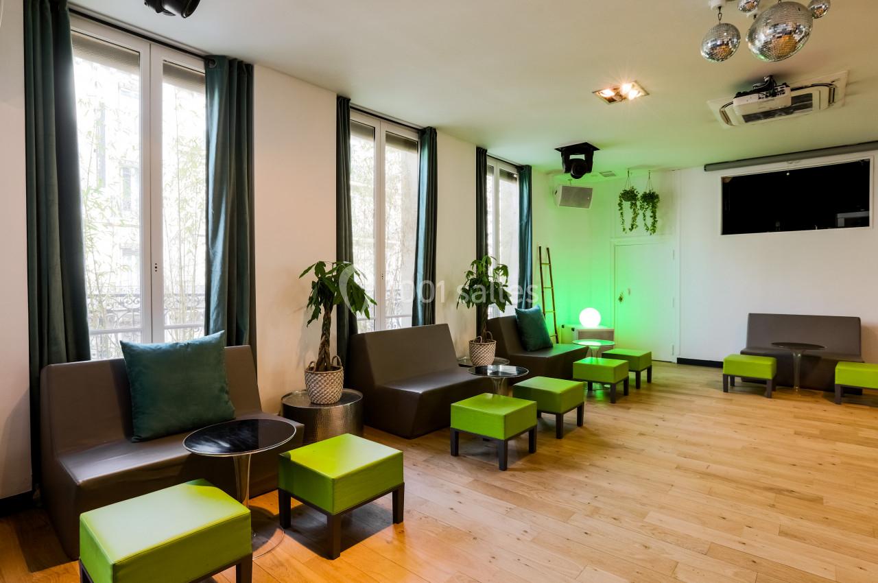 Salle lumineuse avec parquet, banquettes grises, poufs verts, plantes et décoration moderne, éclairée par des fenêtres.