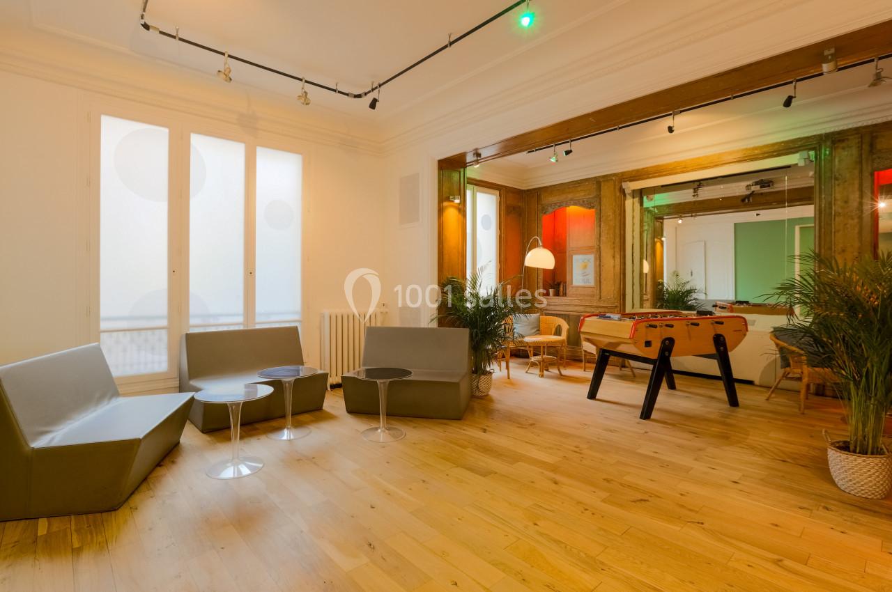 Salle lumineuse avec parquet, canapés gris, baby-foot, plantes et grandes fenêtres laissant entrer la lumière naturelle.