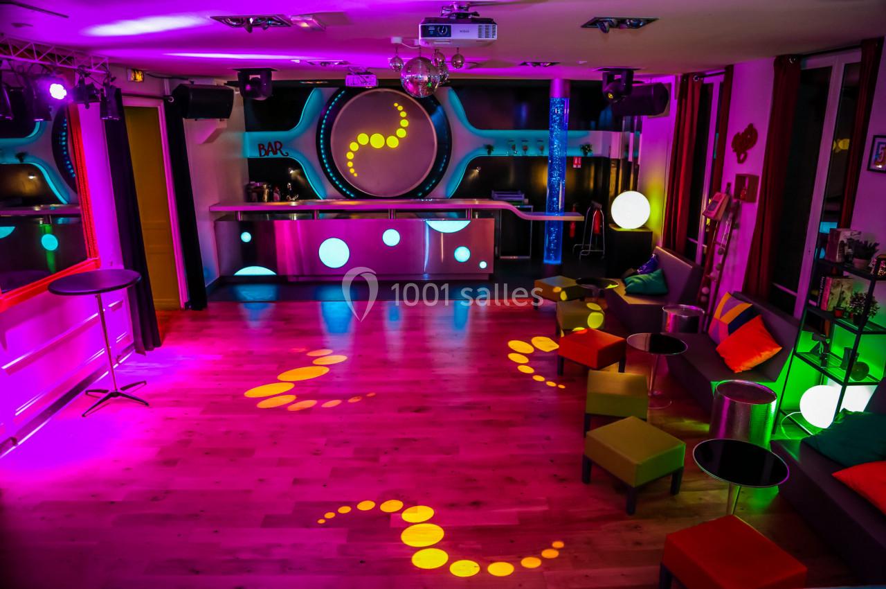 Salle de fête colorée avec éclairages vifs, bar moderne, tabourets et décorations lumineuses sur le sol en bois.
