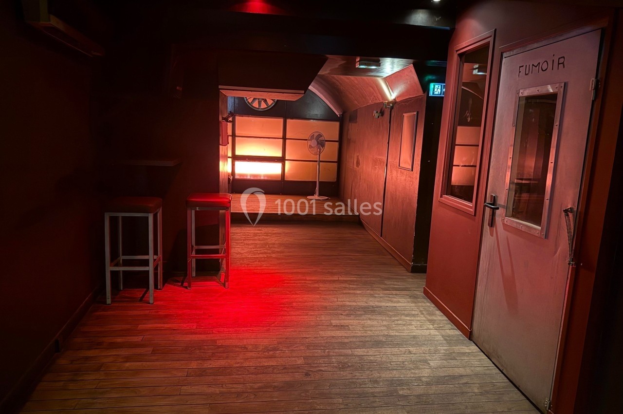 Couloir sombre avec éclairage rouge, sol en bois, porte indiquant ’Fumoir’ et ventilateur visible au fond.
