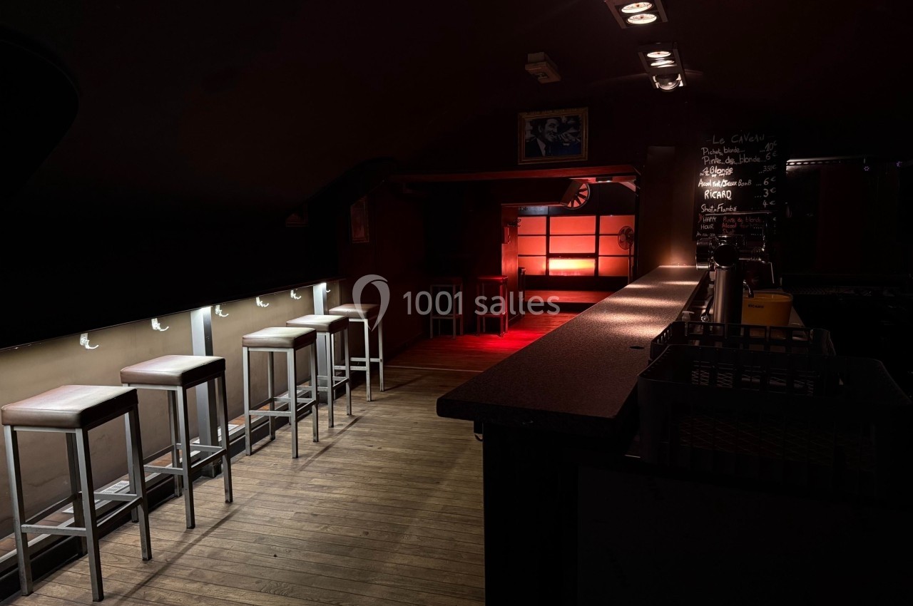 Salle de bar sombre avec un comptoir à droite, des tabourets alignés à gauche et une lumière rouge au fond.