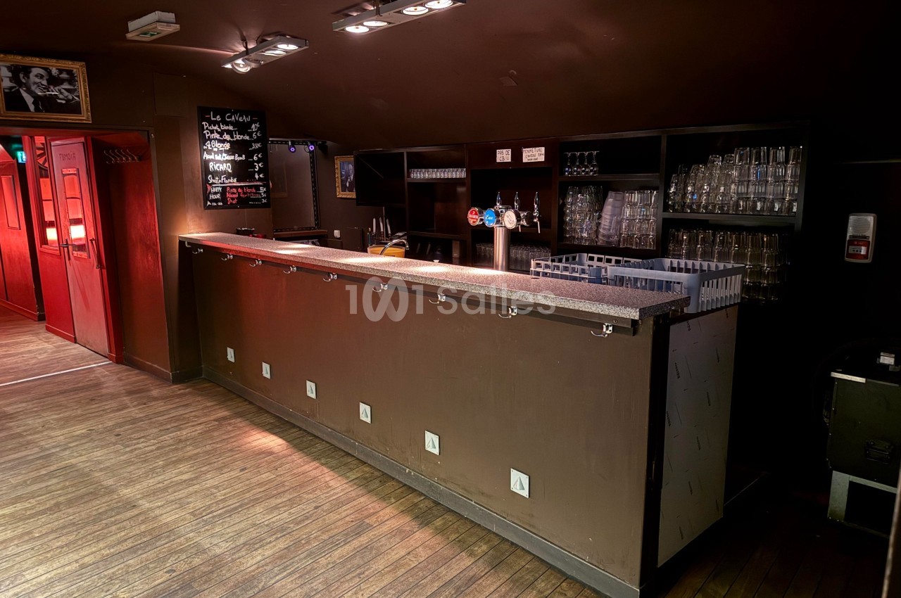 Bar en bois sombre avec comptoir équipé de crochets, étagères à verres et tableau noir affichant le menu.