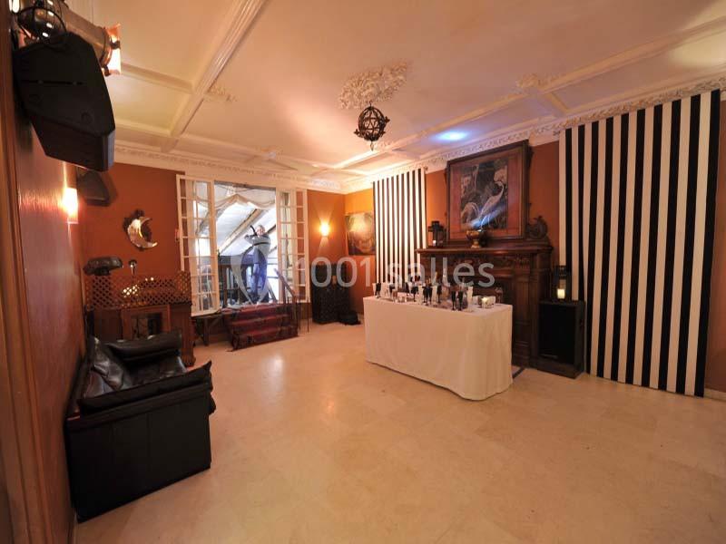 Salle lumineuse avec murs orange, cheminée ornée, table dressée, rideaux rayés noirs et blancs, et grandes fenêtres ouvertes.