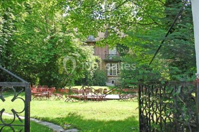 Jardin avec pelouse, tables et chaises rouges, scène en bois, entouré d'arbres et d'une maison à colombages.