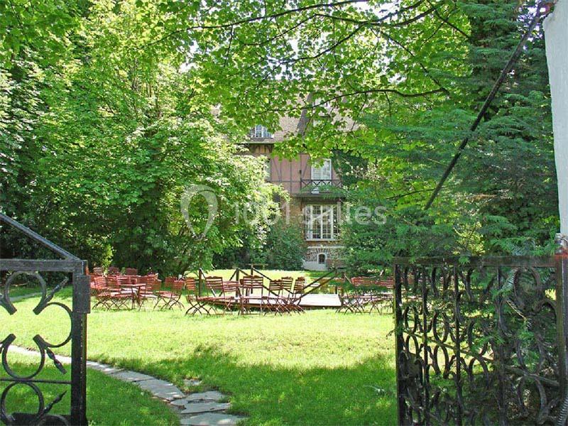 Jardin verdoyant avec des chaises et des tables en bois disposées devant une maison en pierre partiellement visible.