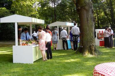 Jardin avec pelouse, tables et chaises rouges, scène en bois, entouré d'arbres et d'une maison à colombages.