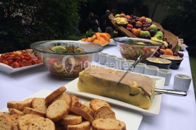 Jardin avec pelouse, tables et chaises rouges, scène en bois, entouré d'arbres et d'une maison à colombages.