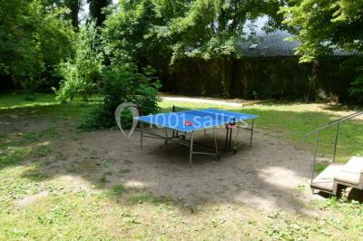 Jardin avec pelouse, tables et chaises rouges, scène en bois, entouré d'arbres et d'une maison à colombages.