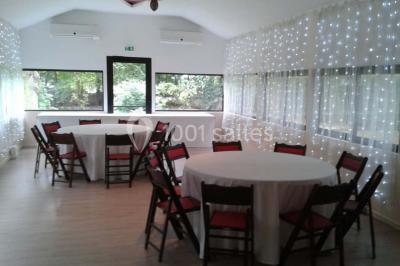 Jardin avec pelouse, tables et chaises rouges, scène en bois, entouré d'arbres et d'une maison à colombages.