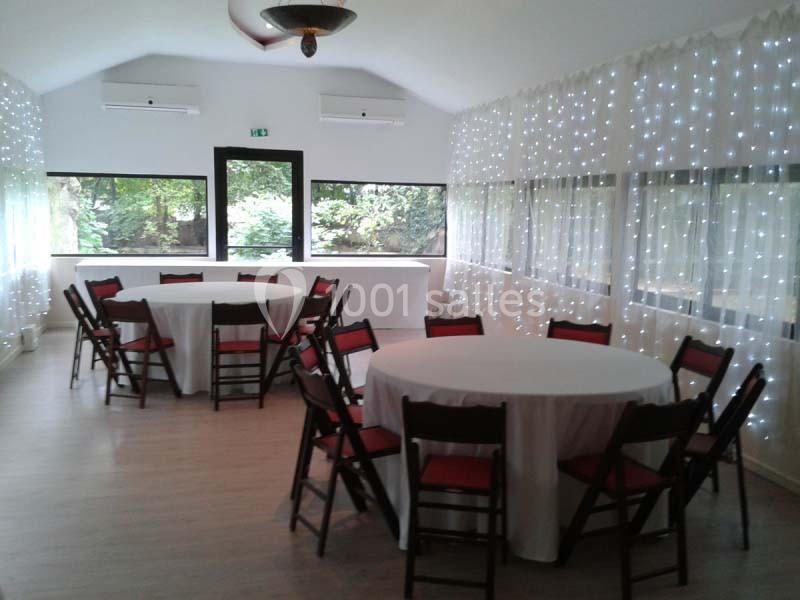 Salle lumineuse avec des tables rondes dressées de nappes blanches, entourées de chaises en bois, et des guirlandes…