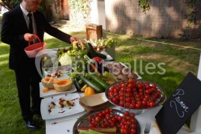 Jardin avec pelouse, tables et chaises rouges, scène en bois, entouré d'arbres et d'une maison à colombages.