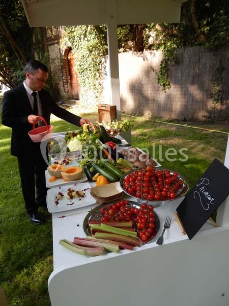 Un homme en costume prépare un buffet de légumes et fruits frais disposés sur une table en extérieur.