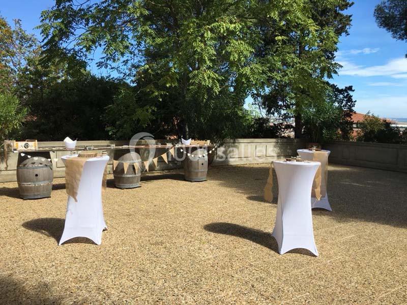 Espace extérieur avec tables hautes drapées de blanc, tonneaux en bois et guirlandes, entouré de verdure.