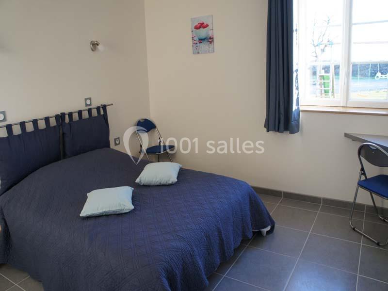 Chambre avec lit double, couvre-lit bleu, deux coussins, bureau, chaise et fenêtre laissant entrer la lumière naturelle.