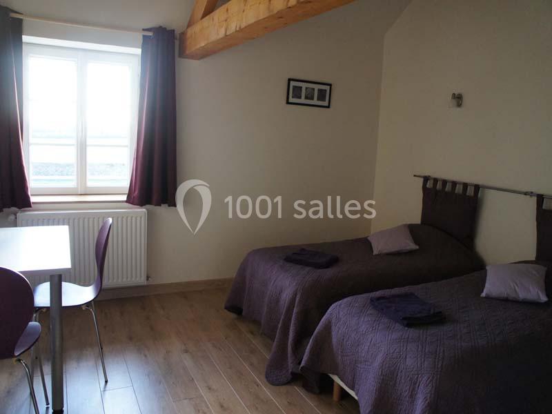 Chambre avec deux lits simples, couvre-lits violets, table et chaises, fenêtre avec rideaux sombres.