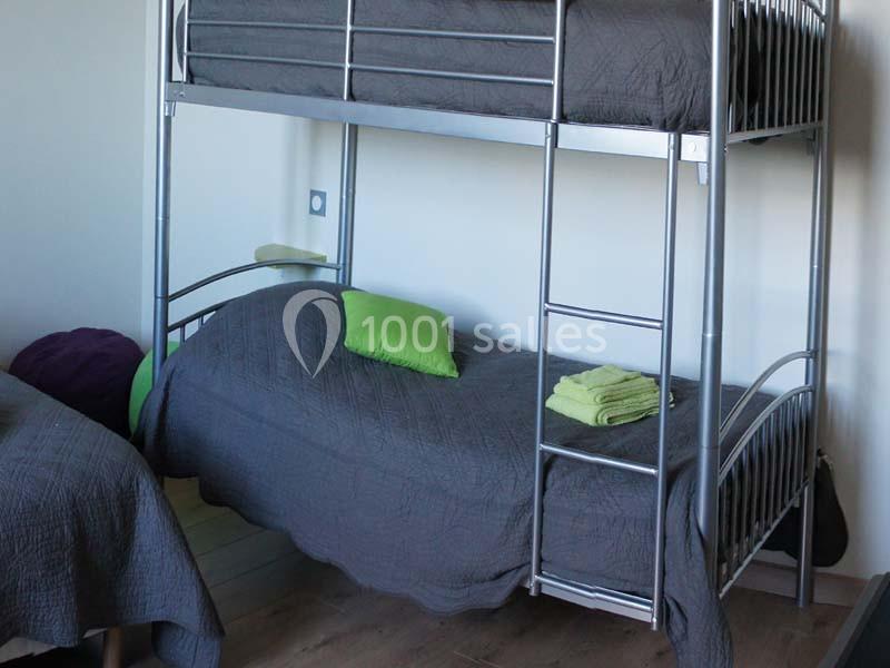 Lit superposé en métal avec draps gris, oreillers verts et serviettes pliées sur le lit inférieur.