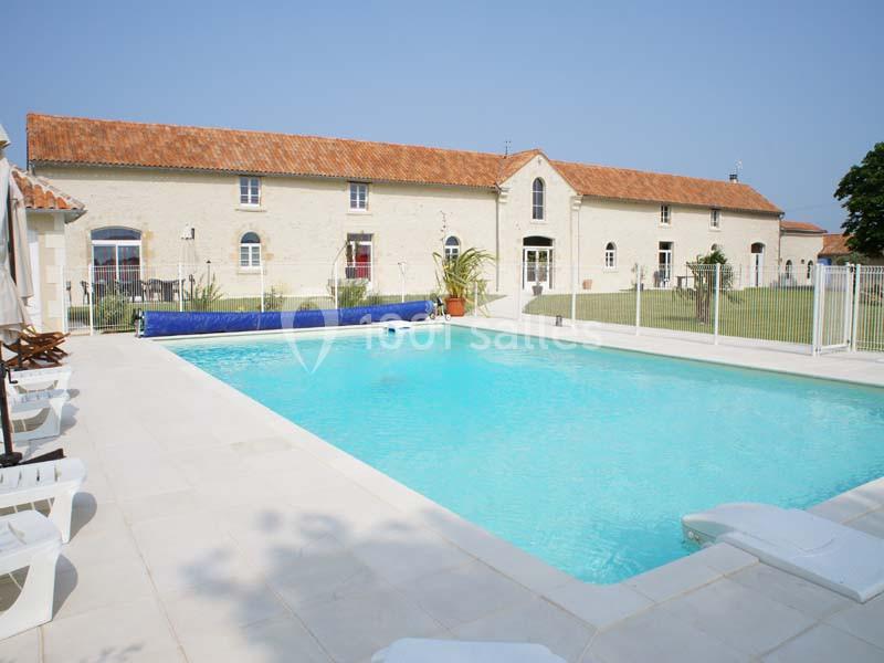 Grande piscine extérieure entourée de chaises longues, située devant un bâtiment en pierre avec toit en tuiles.